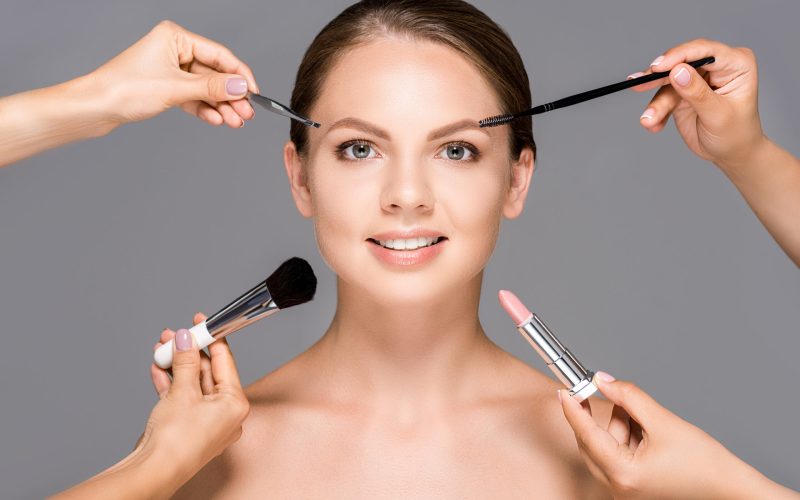 partial view of makeup artists with various cosmetics and smiling woman isolated on grey